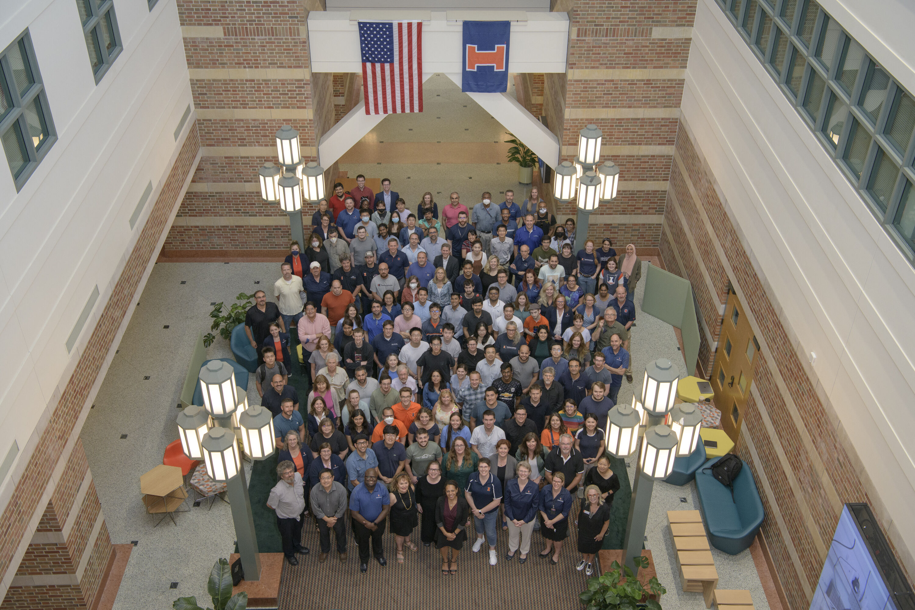 Members of the Beckman Institute community nearly fill the East Atrium as they gather together and smile up at a camera positioned high above.