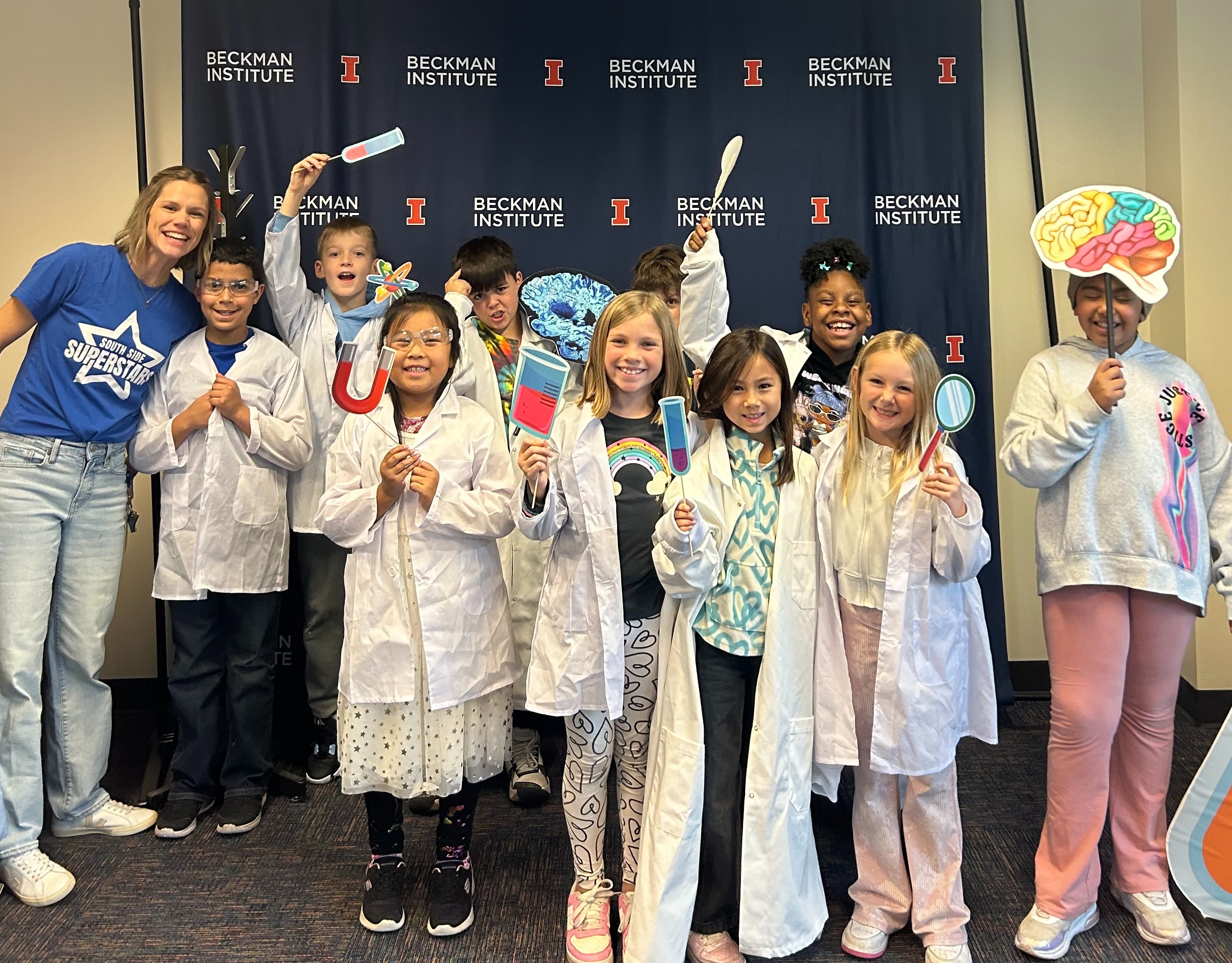 A group of elementary school students poses, dressed up in lab coats and holding paper cutouts of scientific equipment.