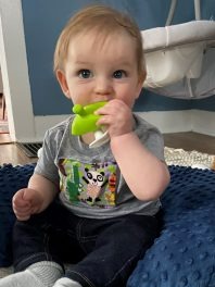 A young child propped up on a pillow, chewing a toy and wearing a LittleBeats shirt with a colorful front pocket.