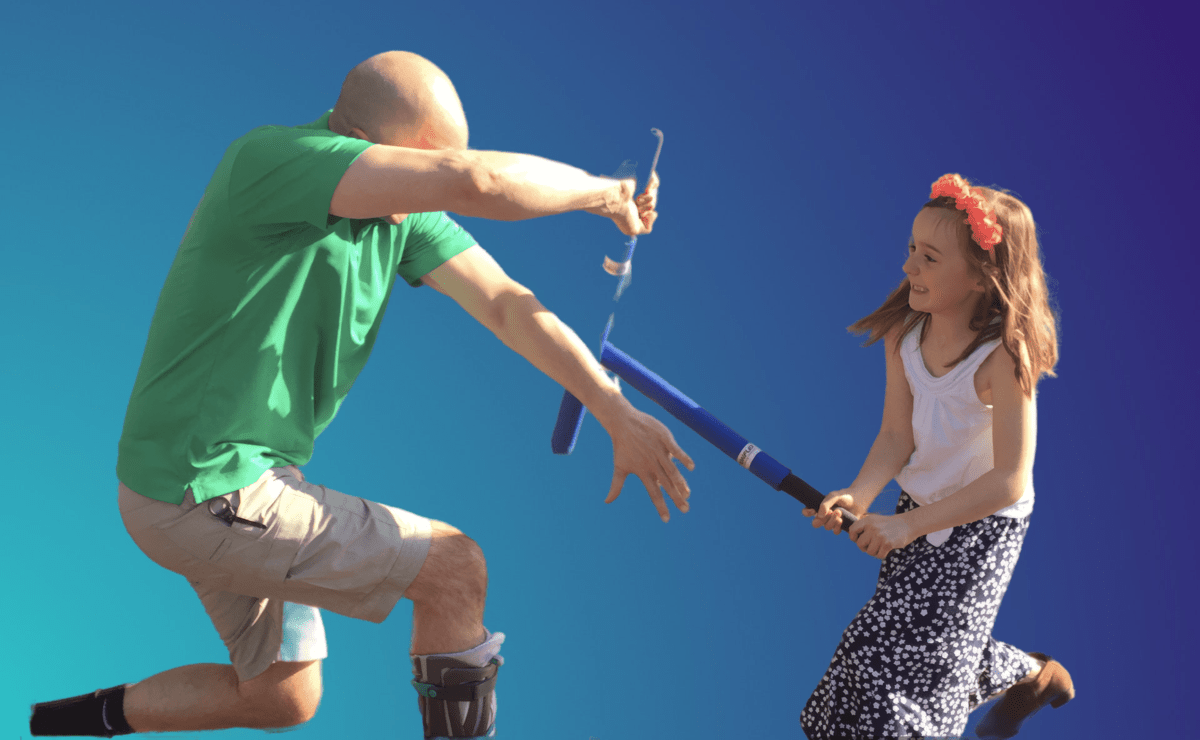 a man in a green shirt and shin guards and a young girl with brown hair and a headband playing with foam swords