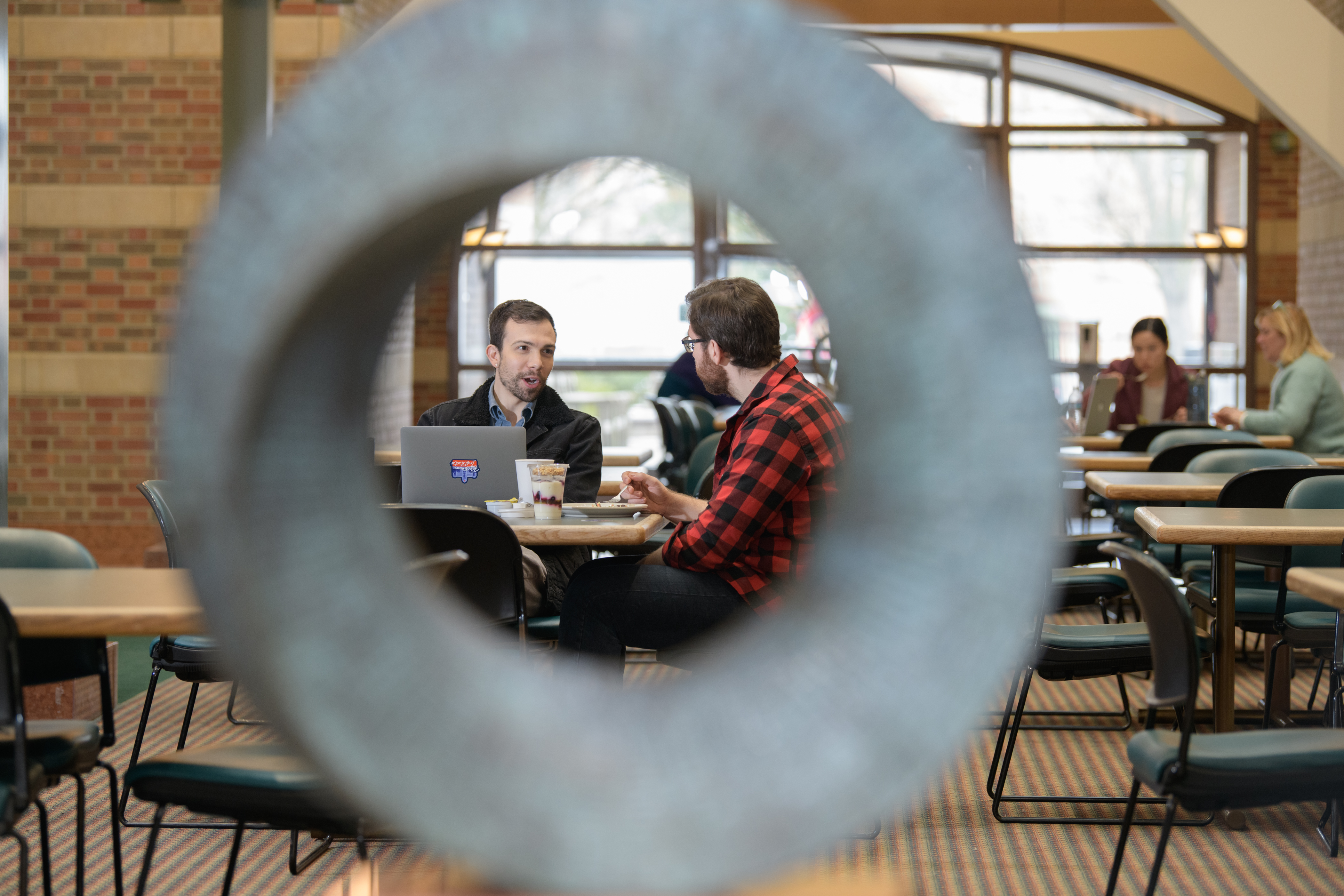 Two Beckman students meet to work and have a bite to eat at the Beckman Café. Captured through the eye of a donut-shaped statue in the Beckman Atrium.