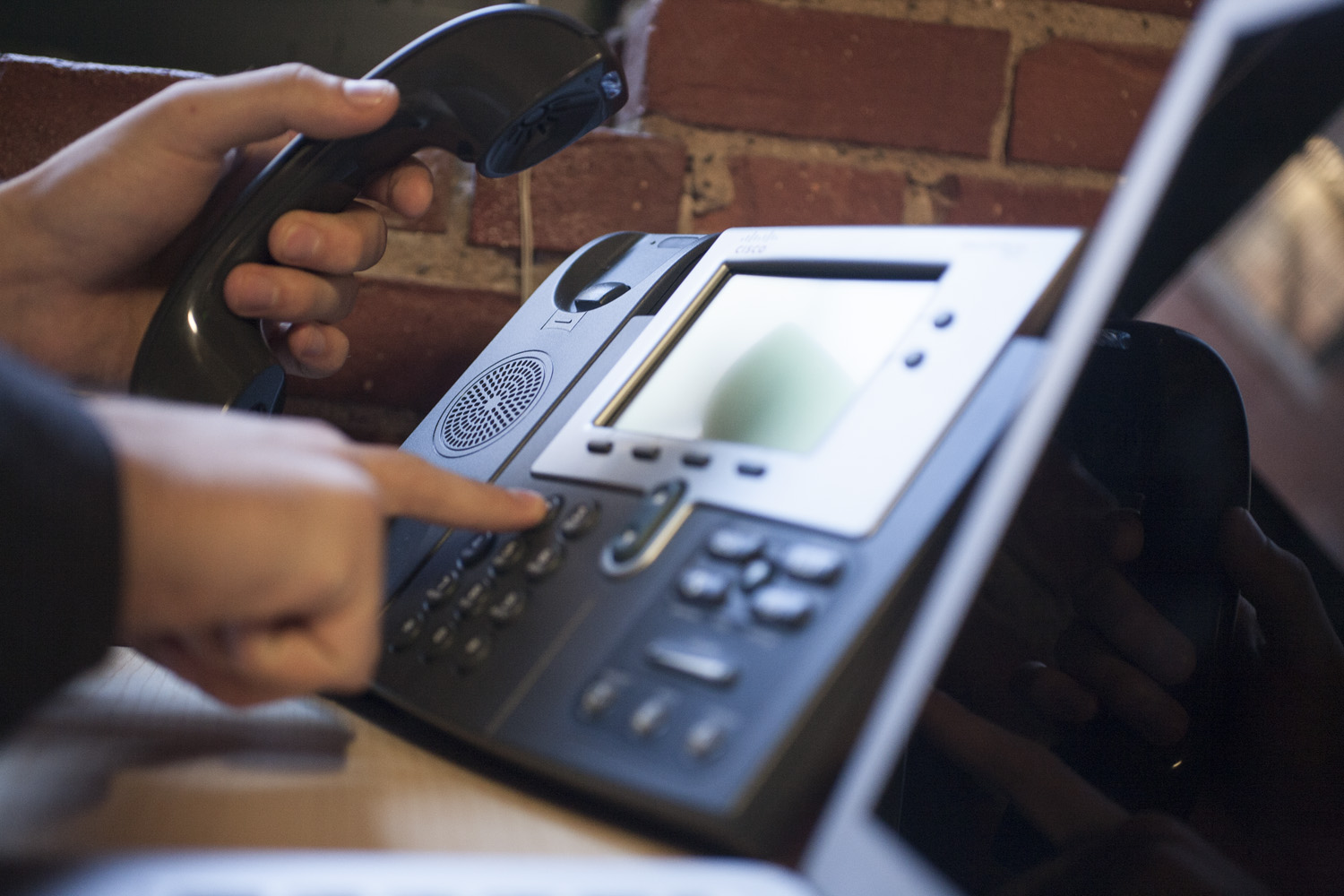A hand dialing an office phone in front of a brick wall.