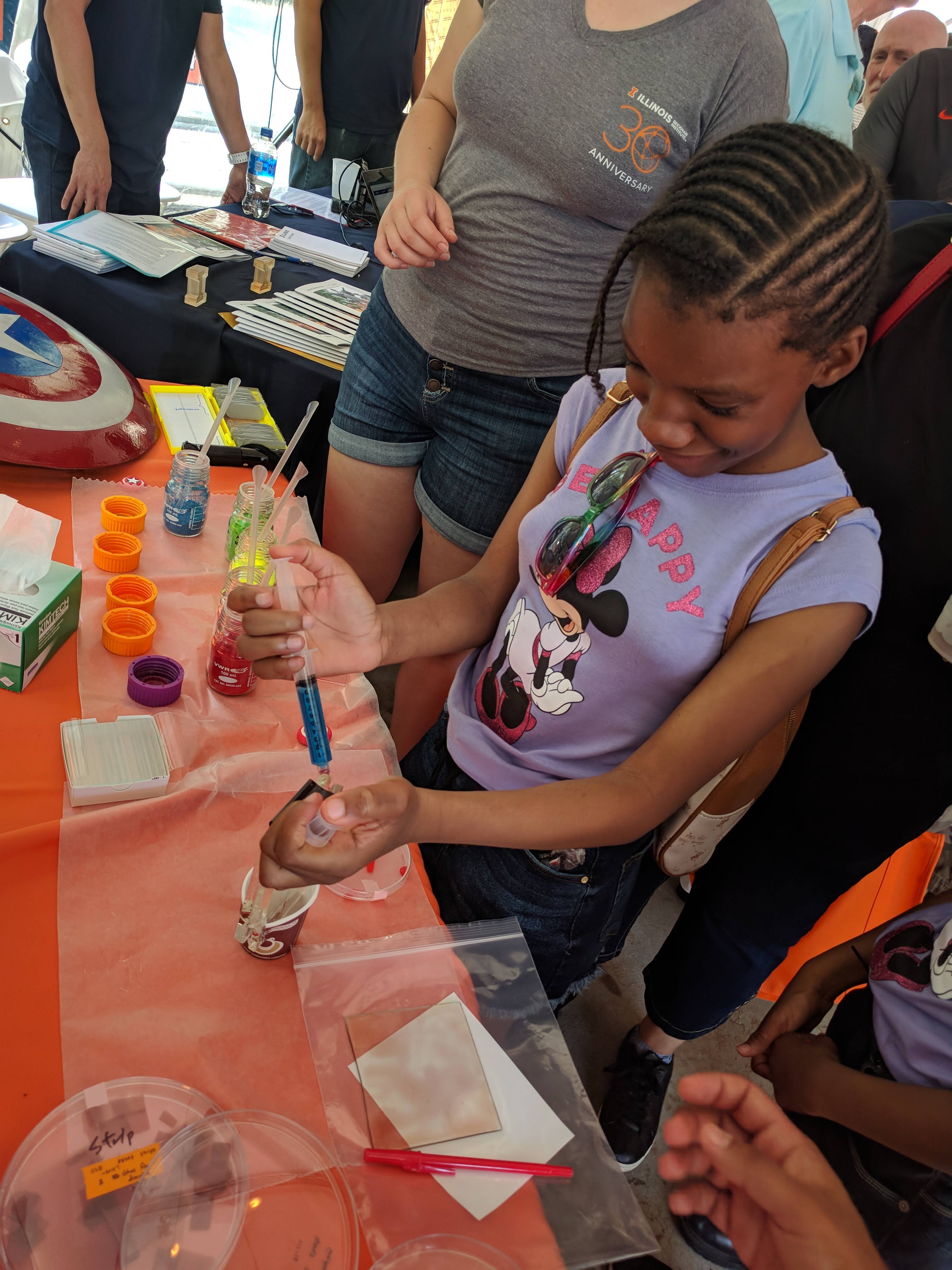 A girl who appears to be in elementary or middle school holds a syringe filled with blue liquid, participating in an interactive experiment with the AMS group.