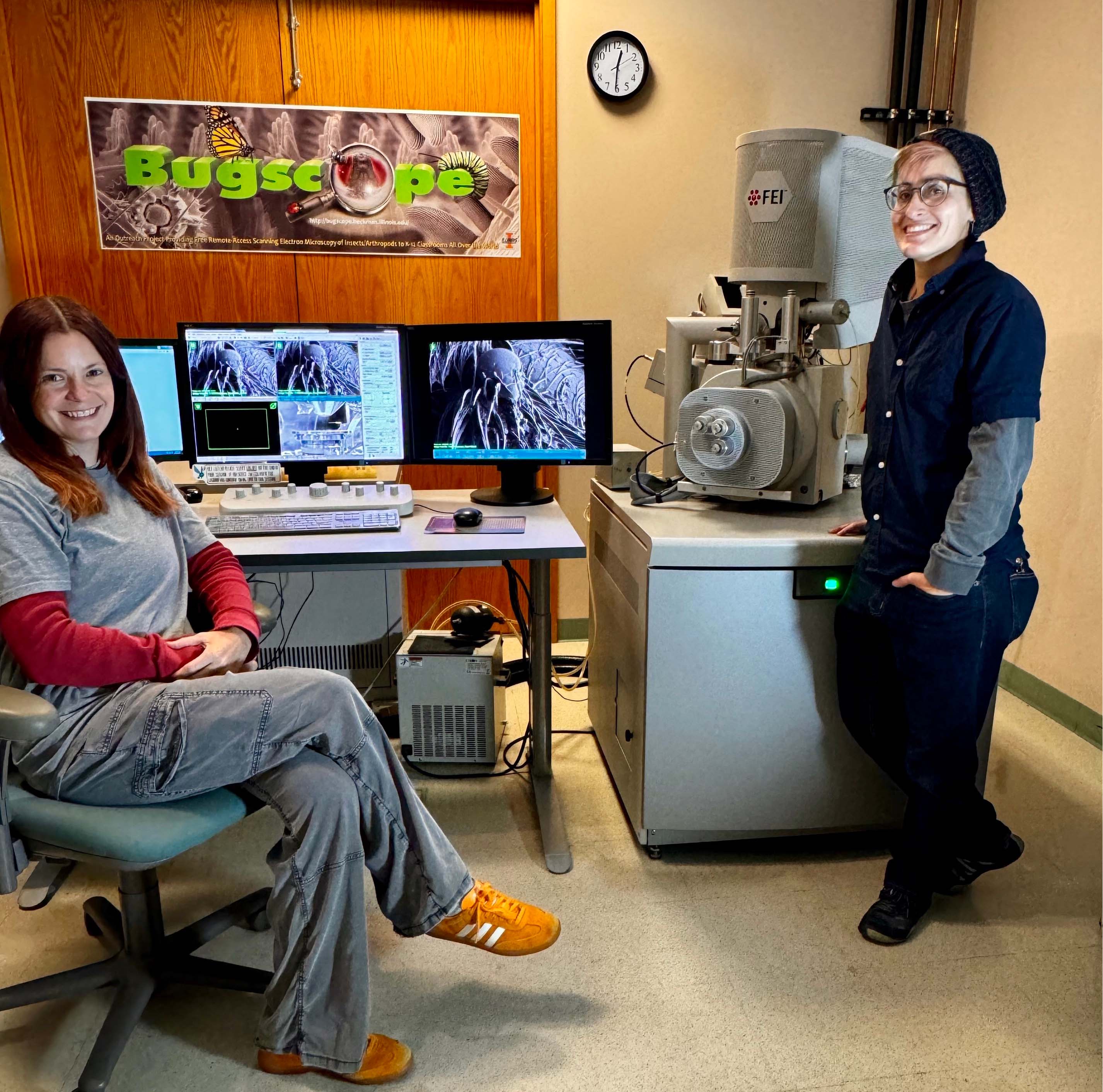 Cate Wallace (left) and T. Josek (right) pose in front of the scanning electron microscope and a Bugscope sign in the Beckman Institute Microscopy Suite.