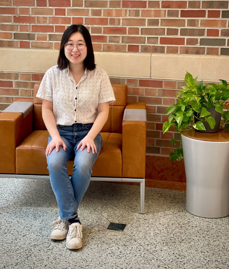 A photograph of Yutao Chen sitting on a leather chair next to a plant in front of a brick wall in the Beckman Atrium