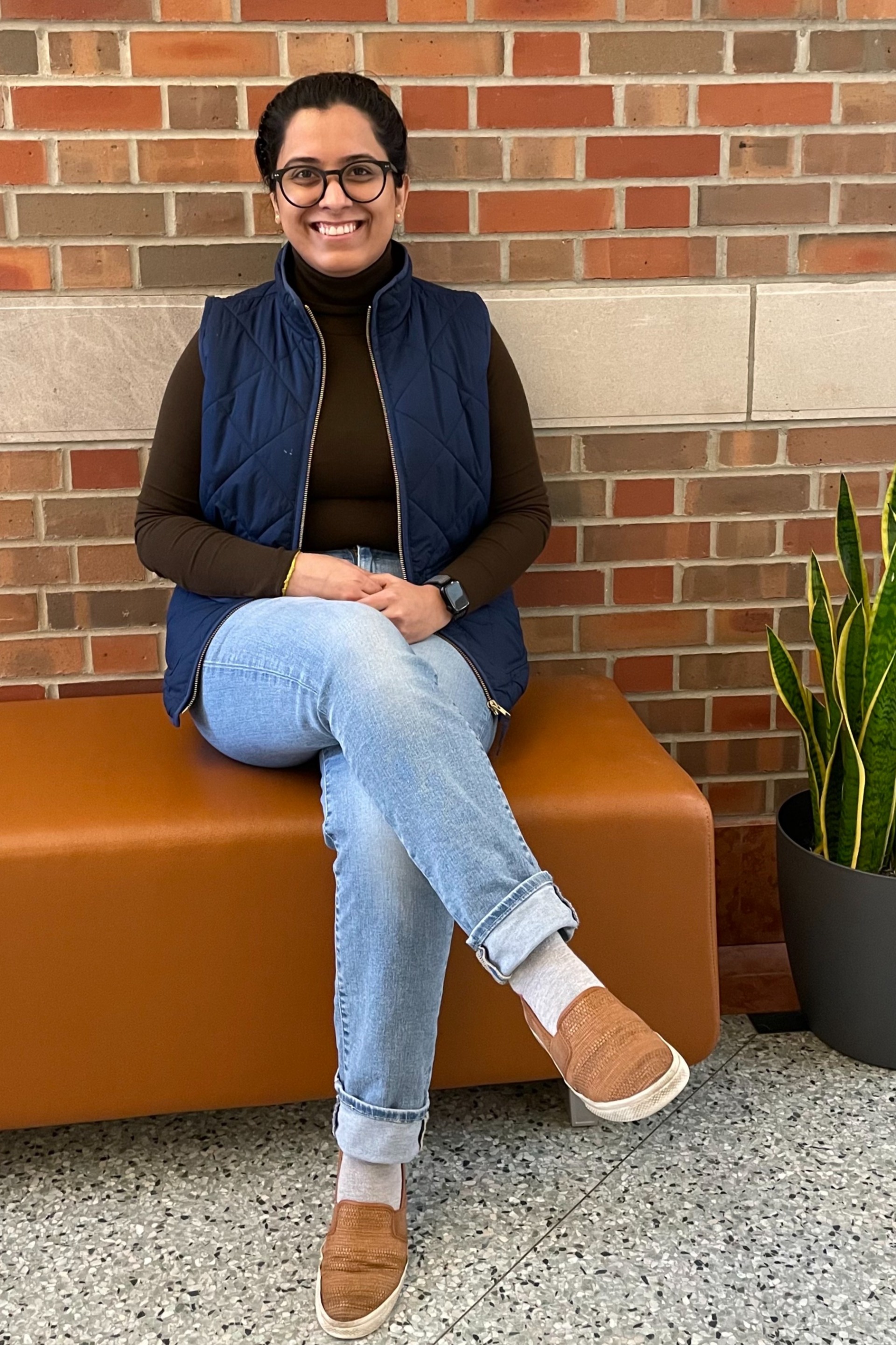 2024 Beckman Postdoctoral Fellow Hemani Chhabra poses on a bench in front of a brick wall inside the Beckman atrium. On her right is a potted snake plant.