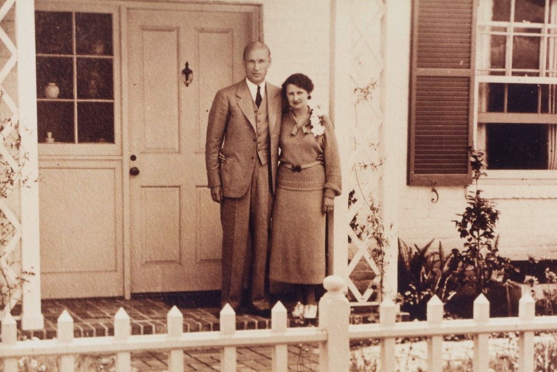 Arnold and Mabel Beckman pose arm-in-arm in front of their home. A white picket fence is visible in the foreground.