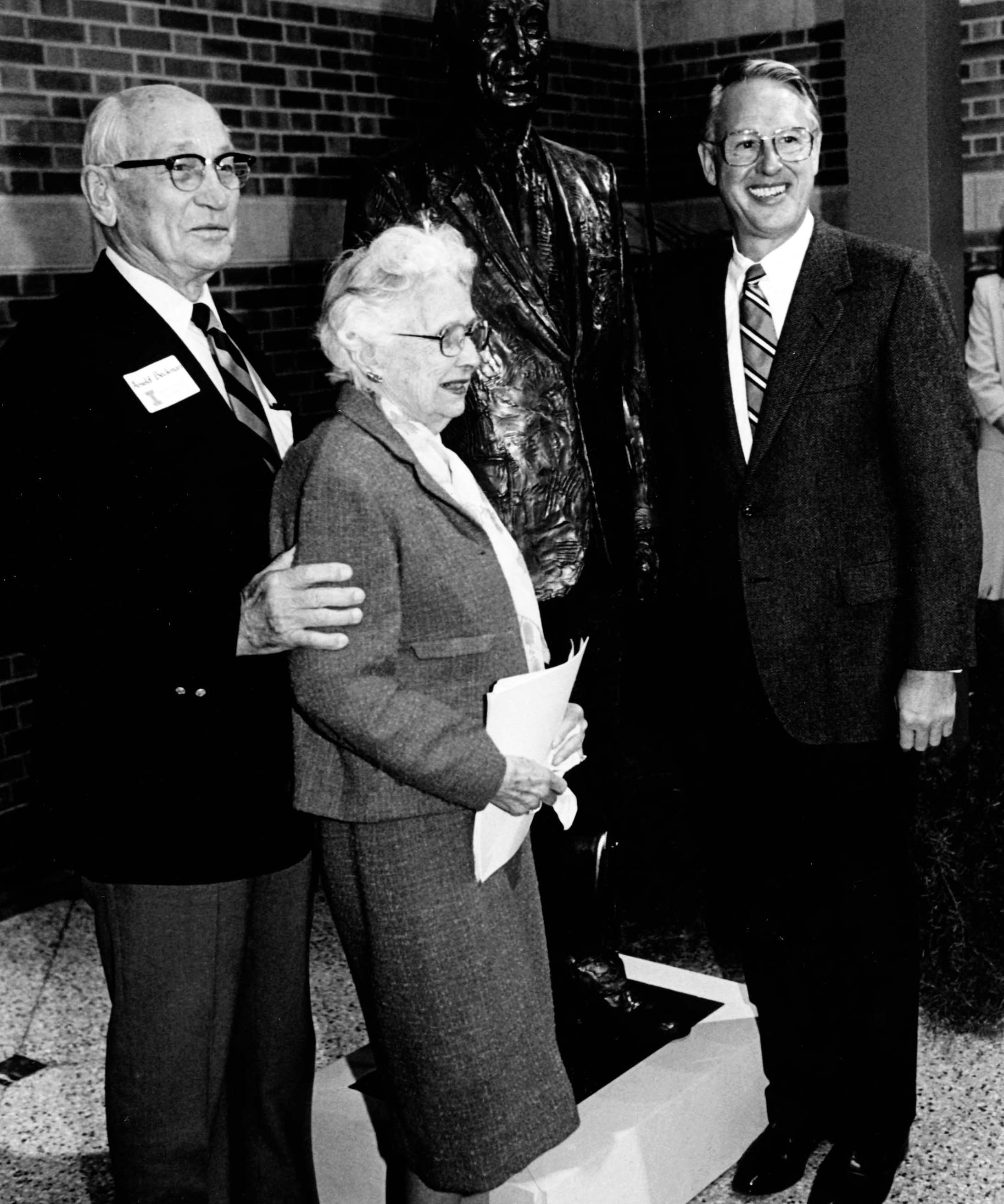 A black-and-white photo depicts Arnold and Mabel Beckman alongside Stanley O. Ikenberry, all posing in front of a bronze statue of Arnold at the Beckman Institute.