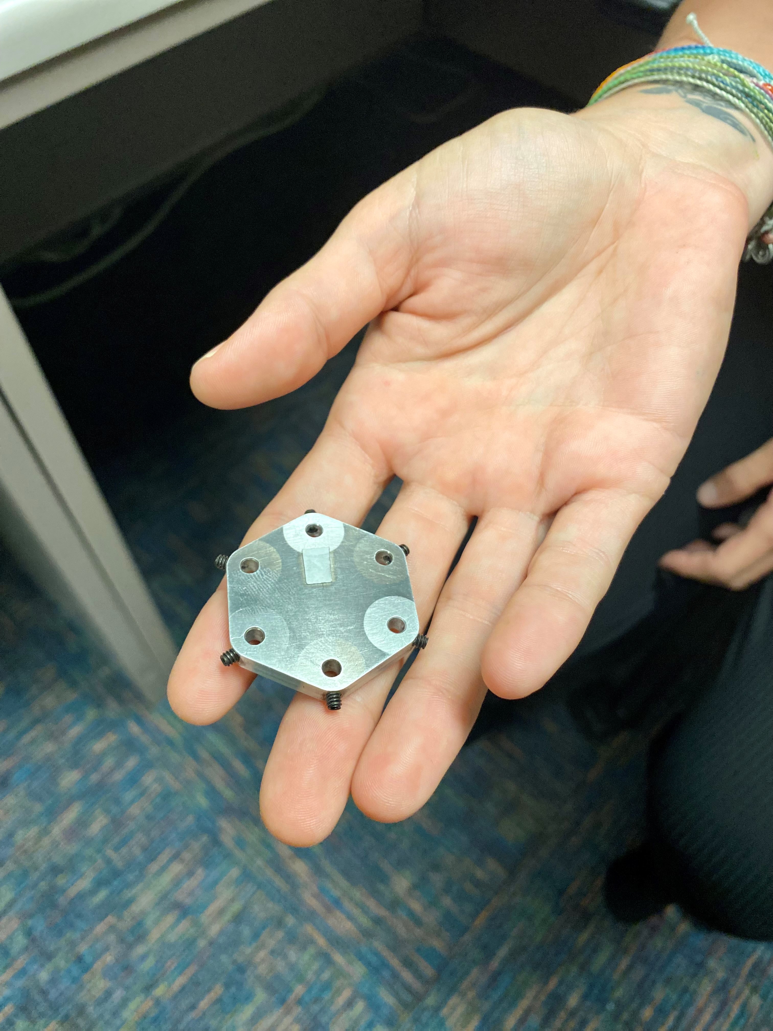 Wallace holds the six-sample holder in her outstretched hand. It is a hexagonal, flat, metal apparatus with six bored holes for storing samples.