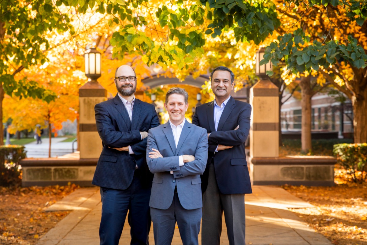 Three male researchers pose outside of the Beckman Institute, illuminated by the brilliant yellow and gold colors of fall leaves.