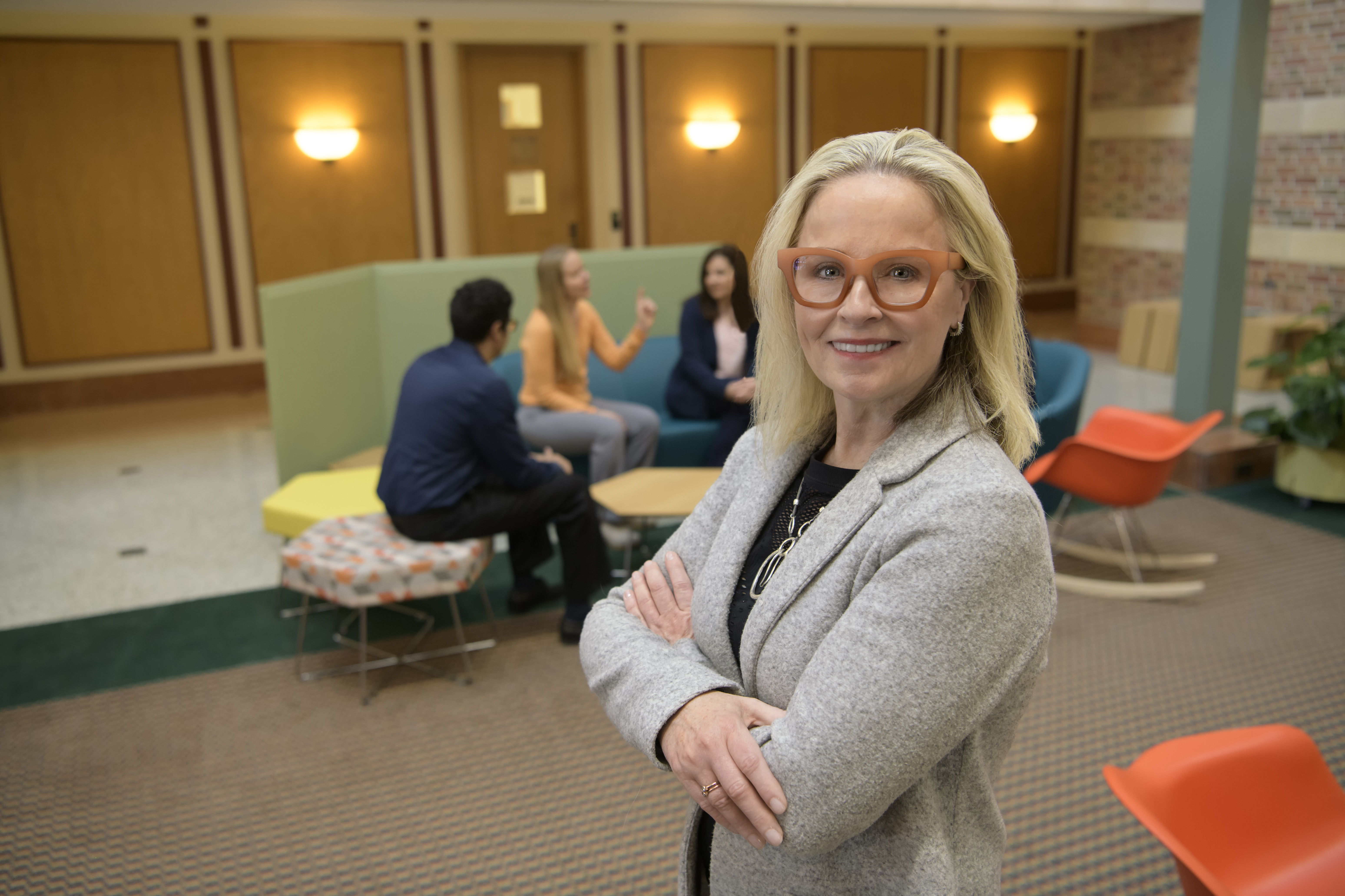 Deana McDonagh poses in the Beckman Atrium; behind her, members of the Beckman community enjoy the furniture she designed for the Beckman Atrium.