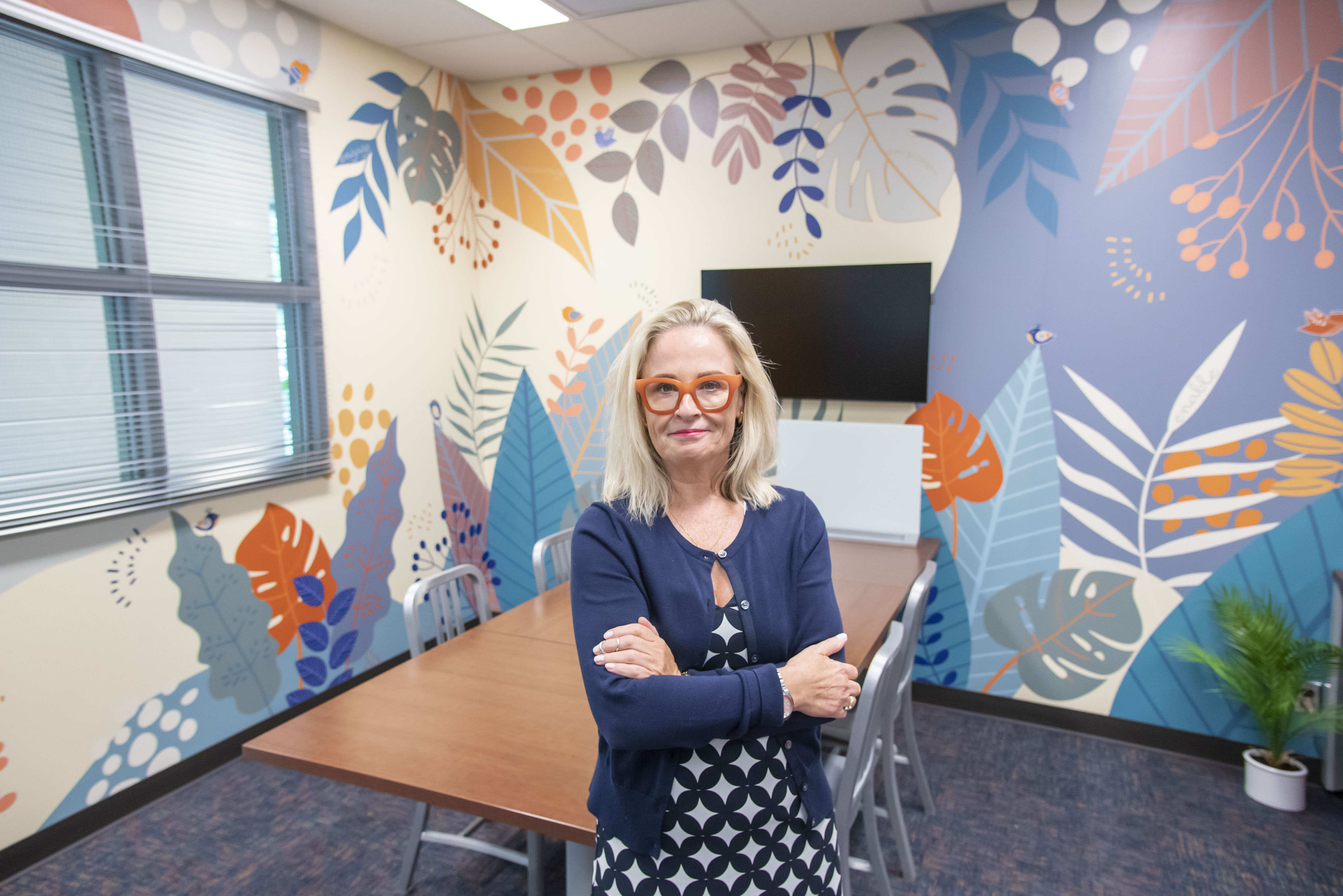 Deana McDonagh stands in the (dis)Ability Design Studio, a colorful mural creating a backdrop behind her.