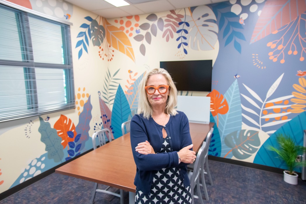 Deana McDonagh stands in the (dis)Ability Design Studio, a colorful mural creating a backdrop behind her.