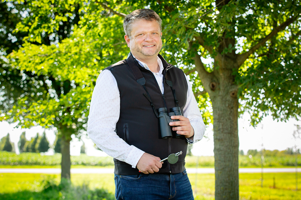 Mark Hauber poses beside a tree outside, holding a pair of binoculars in his hand.
