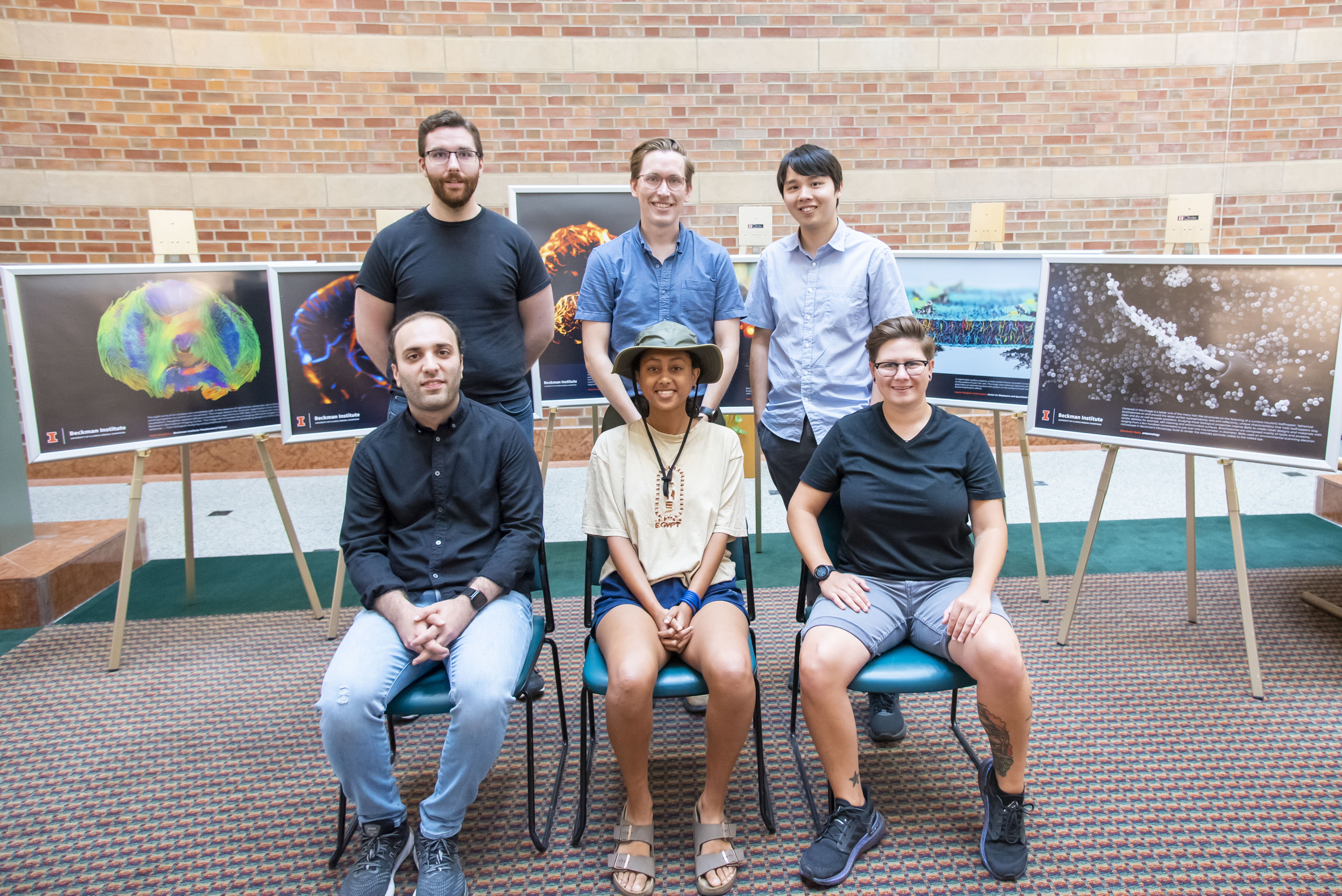 The six winners of the 2022 Beckman Institute Research Image Contest pose, three sitting on chairs and three standing behind them, in front of their winning images, which are framed and mounted on easels.