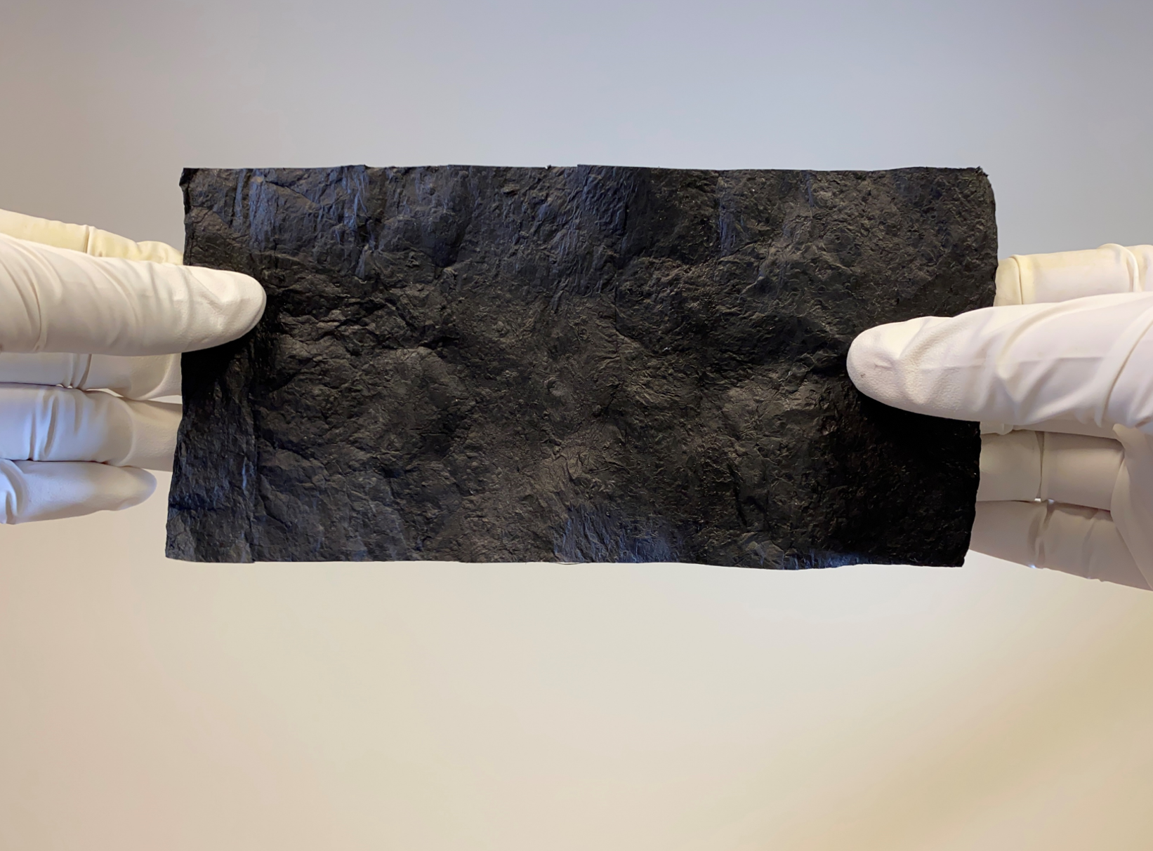 Gloved hands display a carbon nanotube film, which is rectangular and similar in size to a small piece of cardstock.