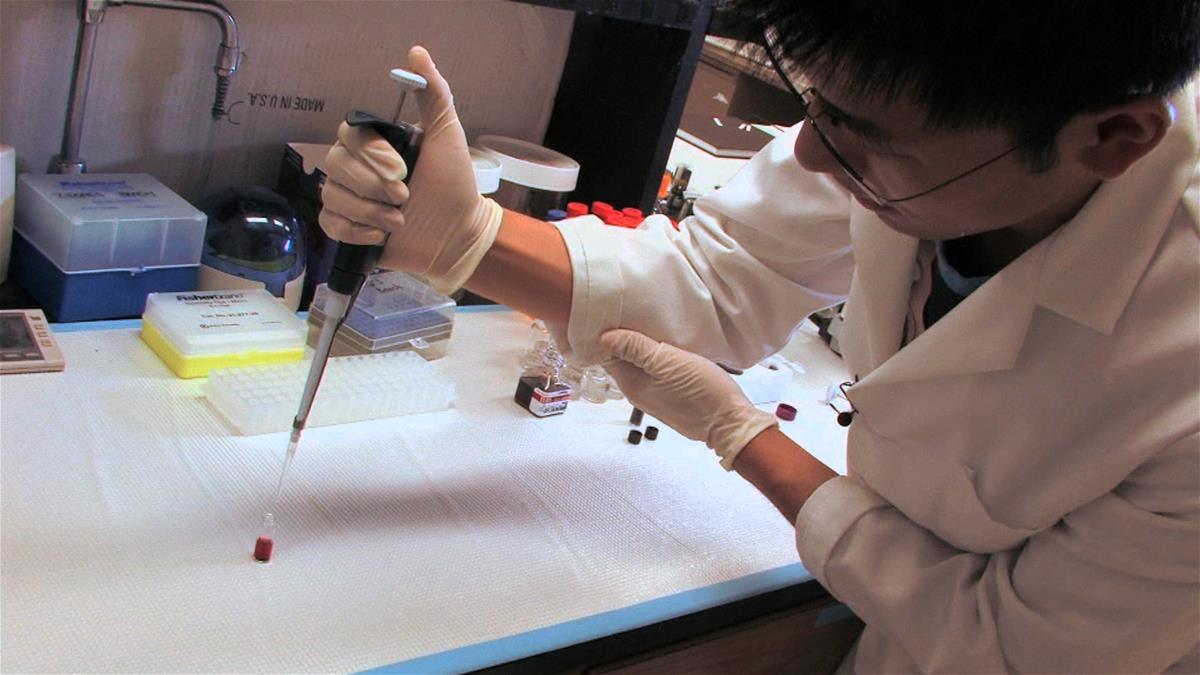A researcher, wearing a lab coat and gloves, holds a pipette over a vial of burgundy liquid in a lab in the Beckman Institute.